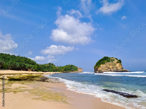 View of beautiful beach with white sand, rocks, green foliages and calm waves under clear blue sky. Summer vacation concept. Ngudel Beach, Malang, Indonesia