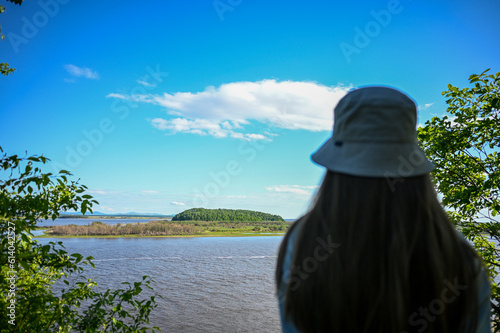 The girl looks into the distance. The river bank. The girl is standing under a big tree. Rest.