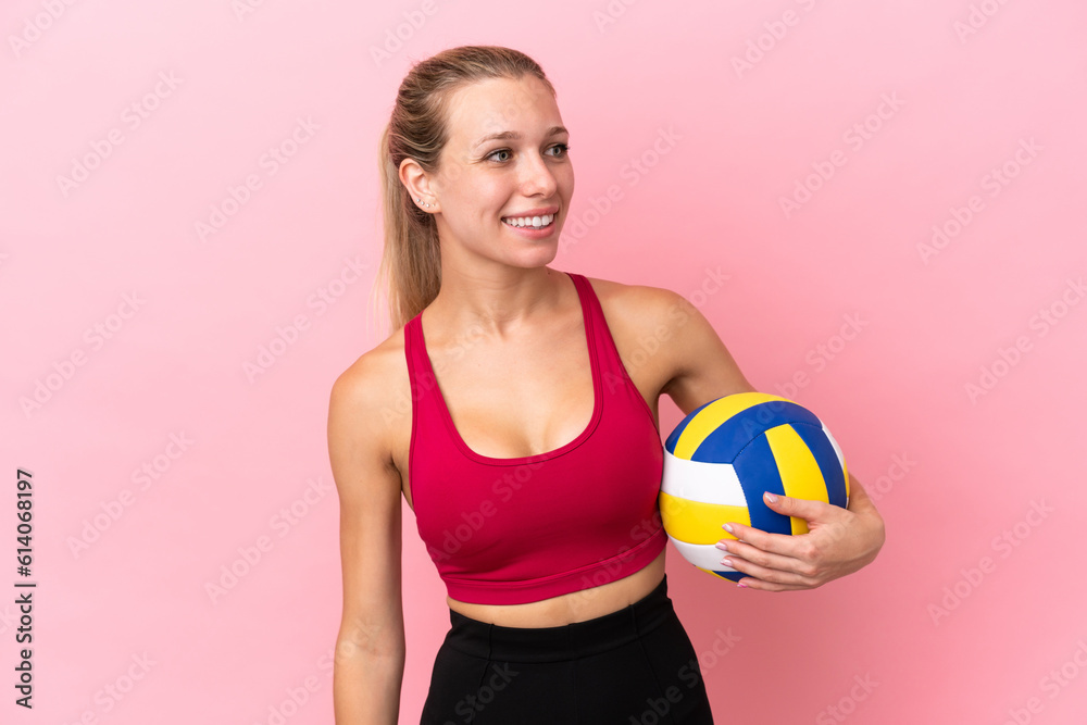 Young caucasian woman playing volleyball isolated on pink background looking to the side and smiling
