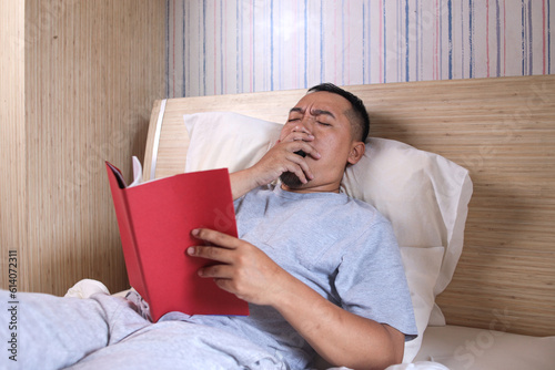 Asian Guy yawning while reading book lying in comfortable bed in bedroom at home. photo