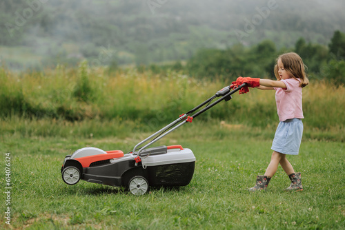 A child in boots in the form of a game mows grass with a lawnmower in the yard against the background of mountains and fog, the concept of garden tools
