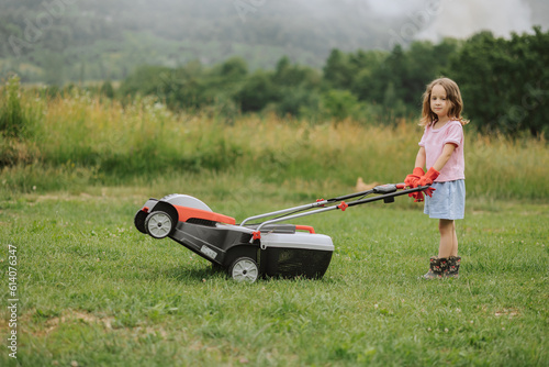 A child in boots in the form of a game mows grass with a lawnmower in the yard against the background of mountains and fog, the concept of garden tools