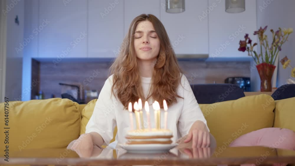 Attractive lonely young woman celebrating birthday, making wish while sitting on yellow sofa and joyfully blowing out candles on cake. girl blows on festive candles at home.