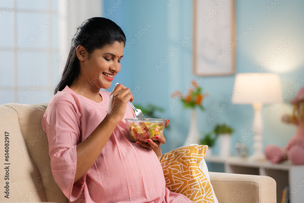 happy-indian-pregnant-woman-eating-fruit-salad-while-sitting-on-sofa-at