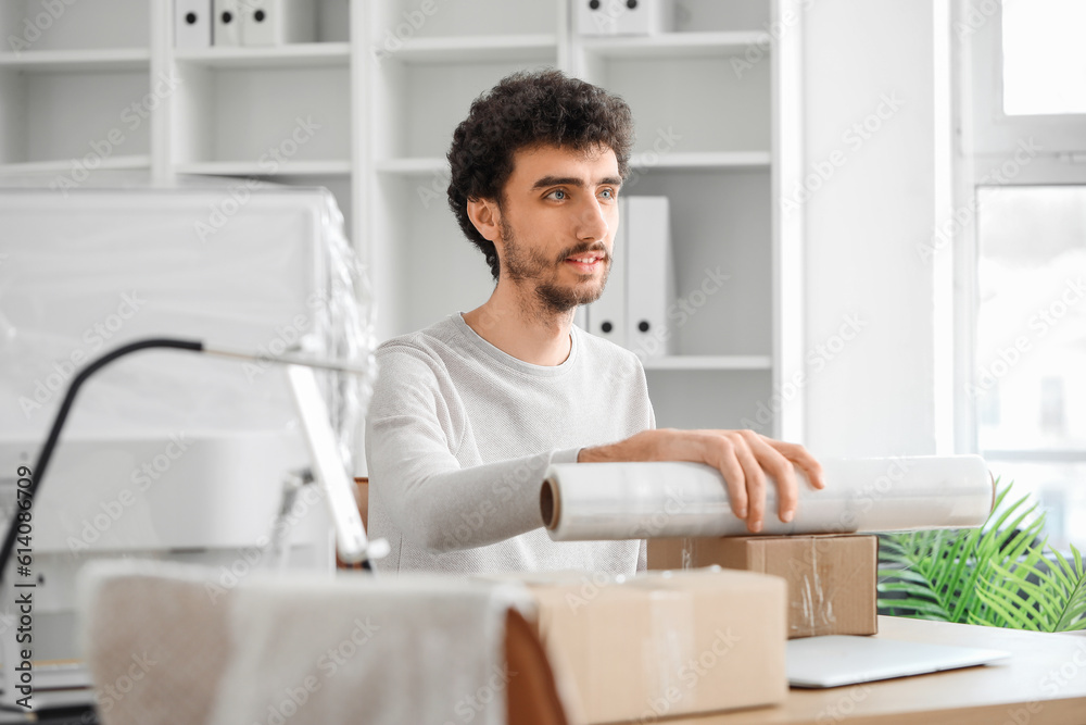 Young man wrapping cardboard box with stretch film in office