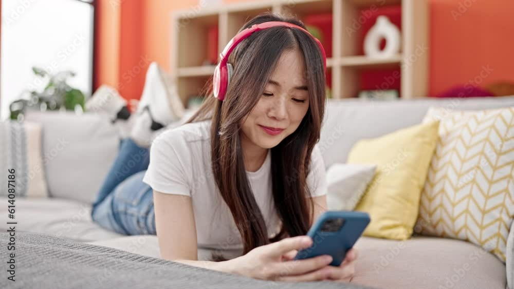 Young chinese woman using smartphone wearing headphones lying on the sofa at home Young chinese woman using smartphone wearing headphones lying on the sofa at home