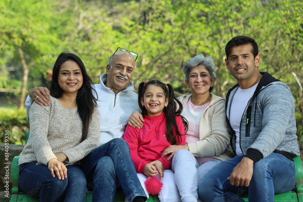 Fototapeta premium Indian happy family having a good cheerful time sitting on a bench in the green park surrounded with a serene atmosphere and fresh air environment in day time.