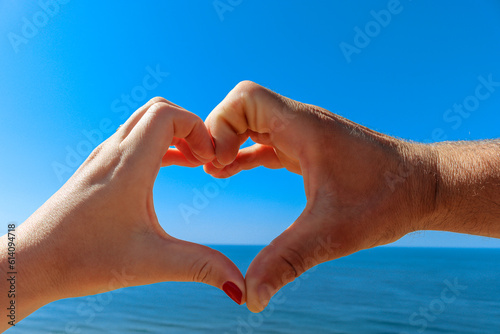 Happy couple holding hand heart gesture with the ocean on background. Young couple holding hands heart-shaped on the sea beach. Complete. © Mihaela