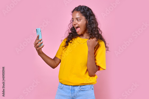 Young overjoyed Indian woman with mobile phone in hands shouts and makes victory gesture after learning about winning jackpot in online casino or stands in pink studio lottery. Betting, excitement