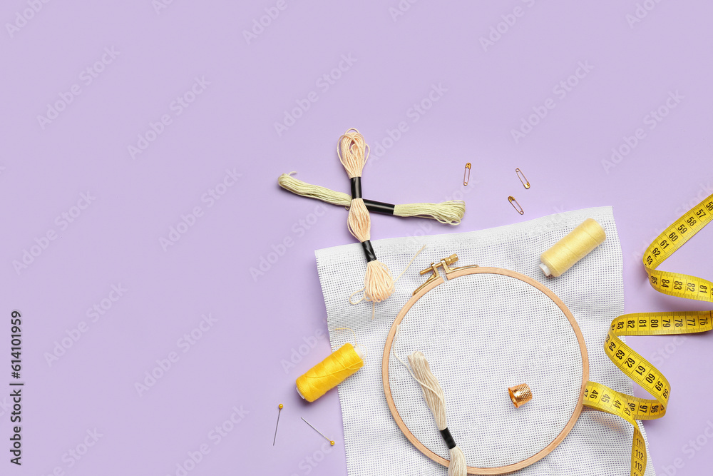 Fototapeta premium Embroidery hoop with canvas, mouline threads and measuring tape on lilac background