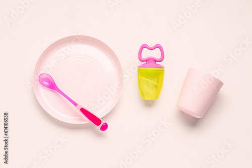 Plate with spoon, cup and nibbler for baby on pink background