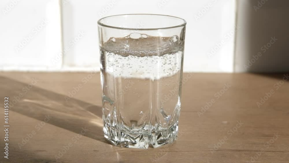 Water pours into transparent glass. Close up pouring purified fresh drink clear water from the bottle on a wooden table in the dining room. Slow Motion of filling up soda pop. Sunlight from the window Water pours into transparent glass. Close up pouring purified fresh drink clear water from the bottle on a wooden table in the dining room. Slow Motion of filling up soda pop. Sunlight from the window