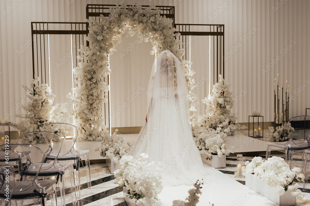Bride in a white elegant wedding long dress walks to the luxury arch decorated with white flowers, back view. Wedding concept.