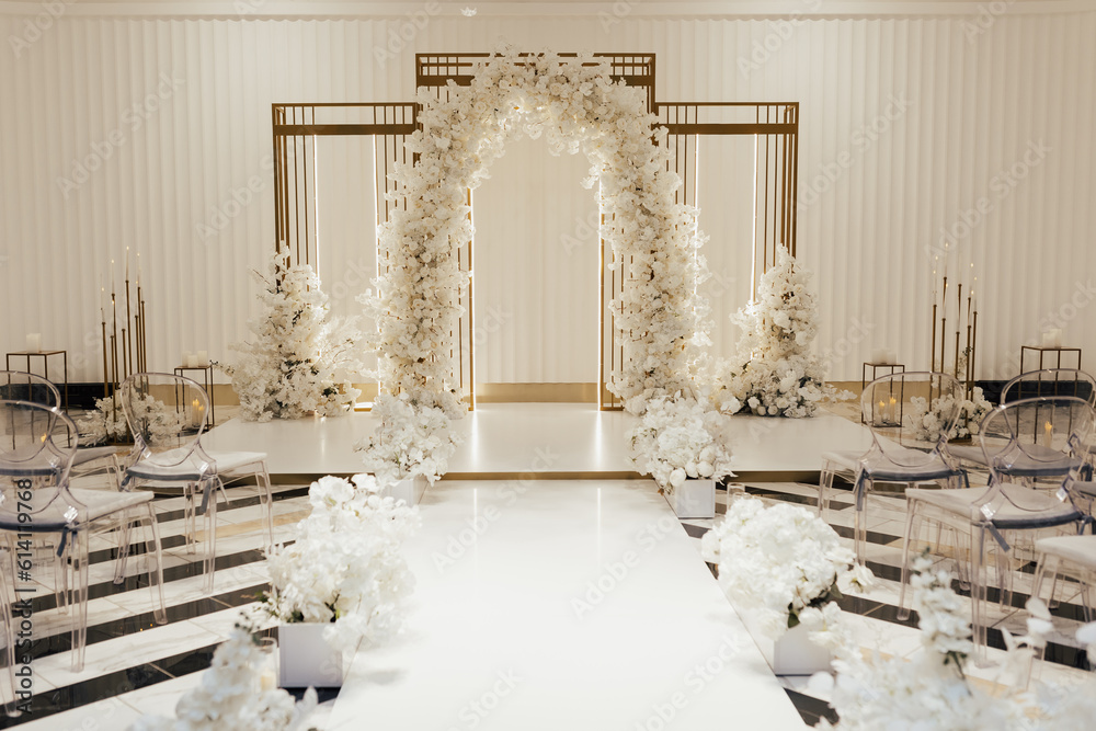 Wedding arch decorated by white flowers standing in the center of wedding ceremony in the luxury restaurant. Transparent chairs on sides.