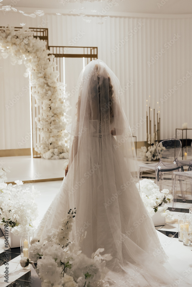 Bride in a white elegant wedding long dress walks to the luxury arch decorated with white flowers, back view. Wedding concept.
