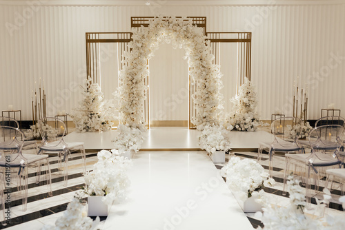 Wedding arch decorated by white flowers standing in the center of wedding ceremony in the luxury restaurant. Transparent chairs on sides. 