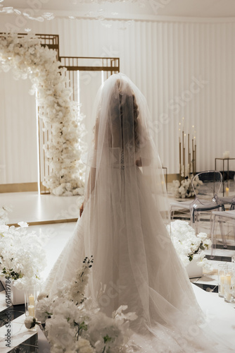 Obraz na płótnie Bride in a white elegant wedding long dress walks to the luxury arch decorated with white flowers, back view. Wedding concept. w salonie