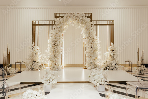 Wedding arch decorated by white flowers standing in the center of wedding ceremony in the luxury restaurant. Transparent chairs on sides.	
 photo