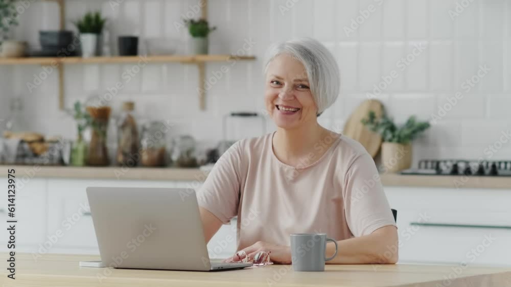 Smiling stylish mature middle aged woman sits at desk with laptop, portrait. Happy senior woman, 60s grey-haired lady wearing glasses looking at camera sitting at home table. Headshot. Smiling stylish mature middle aged woman sits at desk with laptop, portrait. Happy senior woman, 60s grey-haired lady wearing glasses looking at camera sitting at home table. Headshot.