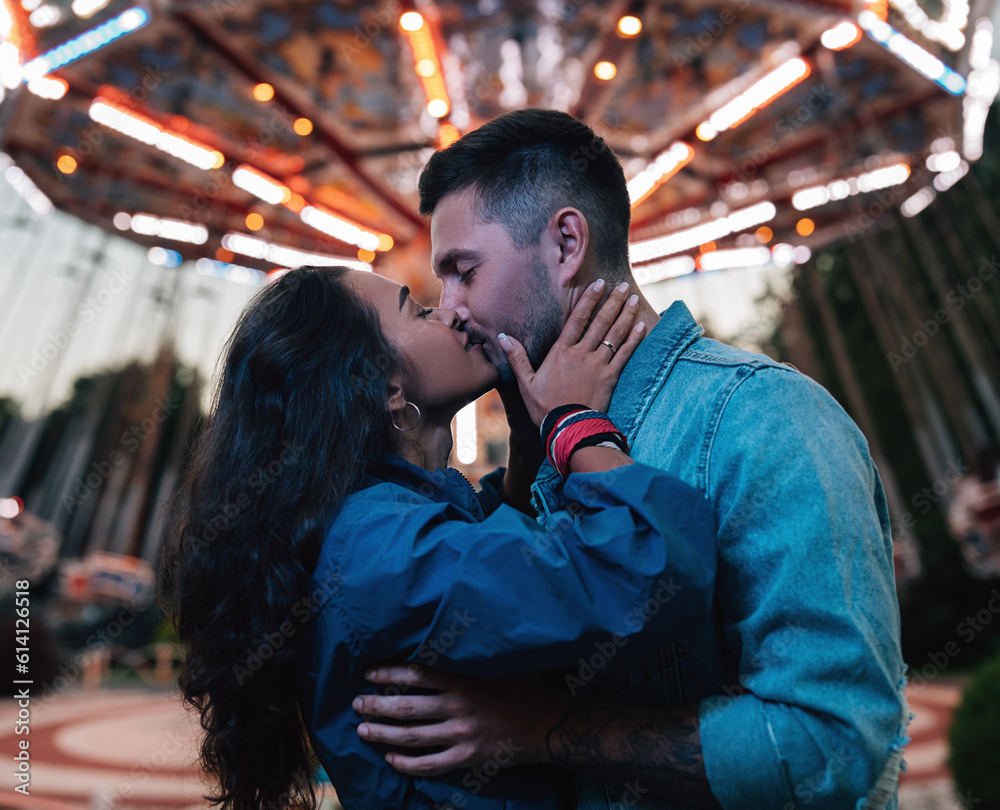 Young couple kissing against carousel. Male and female hugging and kissing in the evening in an amusement park.