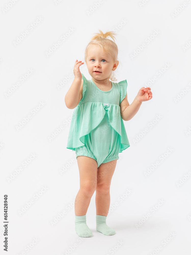 A 2-year-old girl with a questioning expressive look spreads her hands to the side on a white background in a green bodysuit dress. A 2-year-old girl with a questioning expressive look spreads her hands to the side on a white background in a green bodysuit dress.