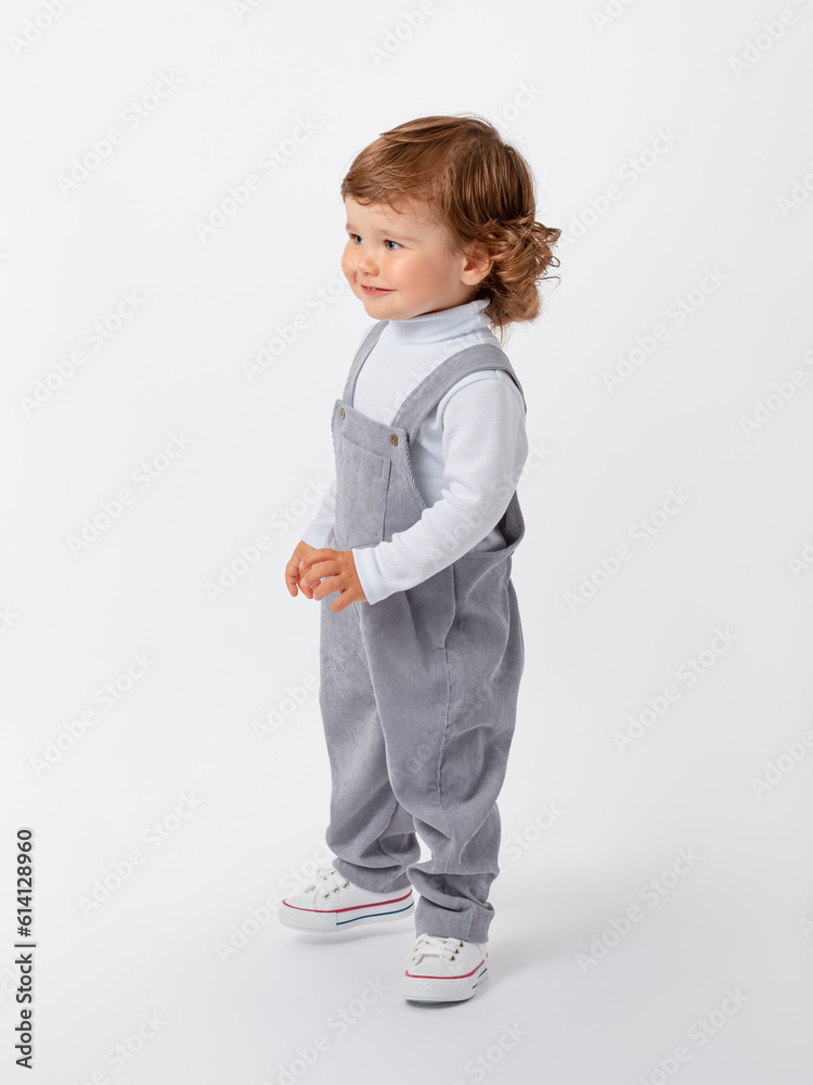 A 2-year-old toddler boy with curly hair is standing sideways, looking out, looking for someone and smiling in a gray jumpsuit and a white turtleneck and sneakers is standing on a white background. A 2-year-old toddler boy with curly hair is standing sideways, looking out, looking for someone and smiling in a gray jumpsuit and a white turtleneck and sneakers is standing on a white background.