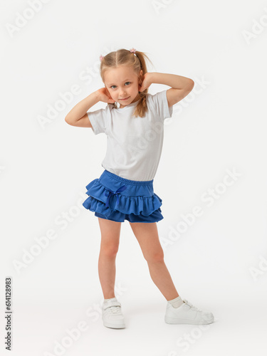 A 5-year-old girl poses with her hands behind her head on a white background and looks expressively at the camera in a white T-shirt, blue skirt and sneakers.
