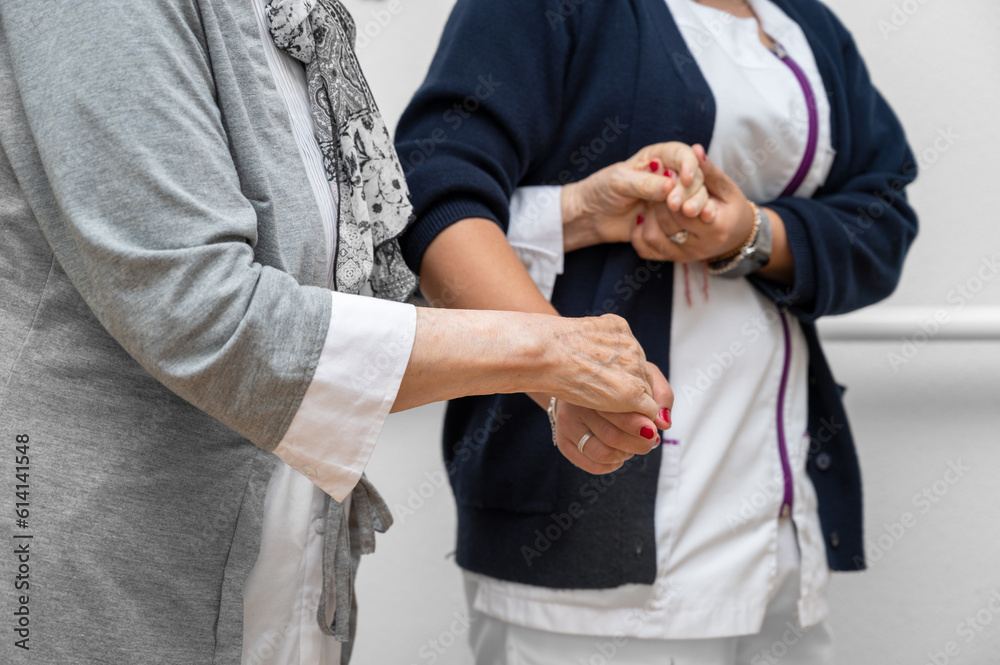 Obraz premium Nurse helping elderly woman walk in the room, holding his hand, supporting her. Treatment and rehabilitation after injury or stroke, life in assisted living facility, senior care concept.