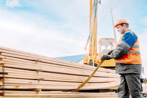 A slinger unloads wooden planks outdoors on a summer day. A worker in a hard hat and high-visibility vest stacks lumber. Industrial background with copy space.