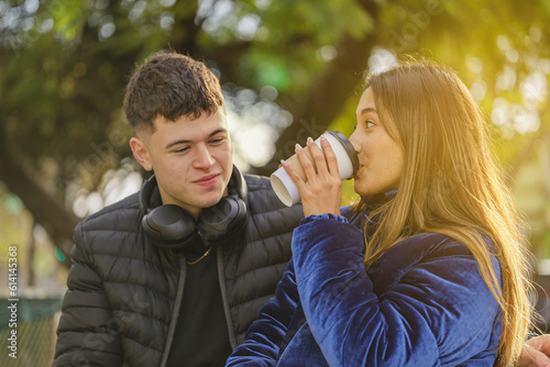 Latina girl drinks coffee in a disposable cup next to a Caucasian boy on the bench in a public park.