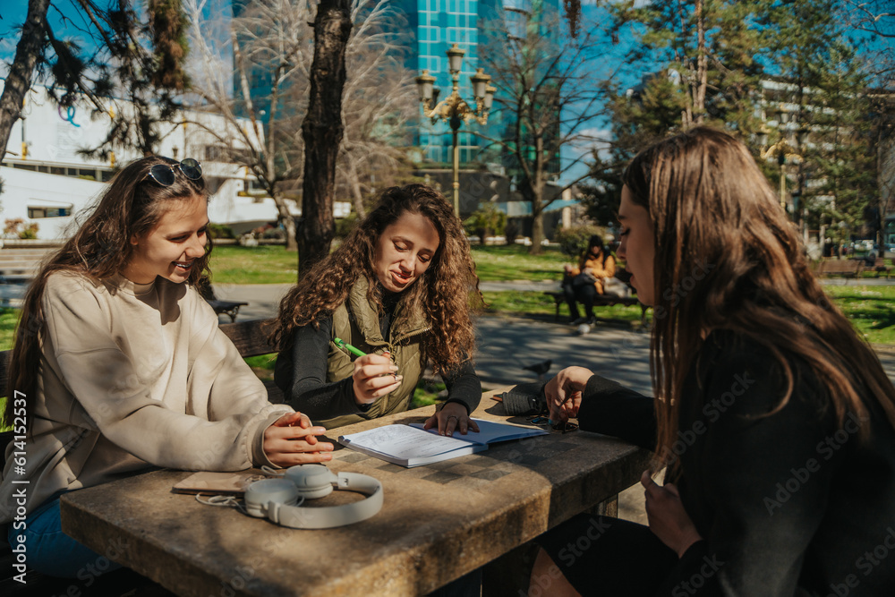 Lovely curly haired girl writing homework while sitting with her friends in the park, on a beautiful sunny day