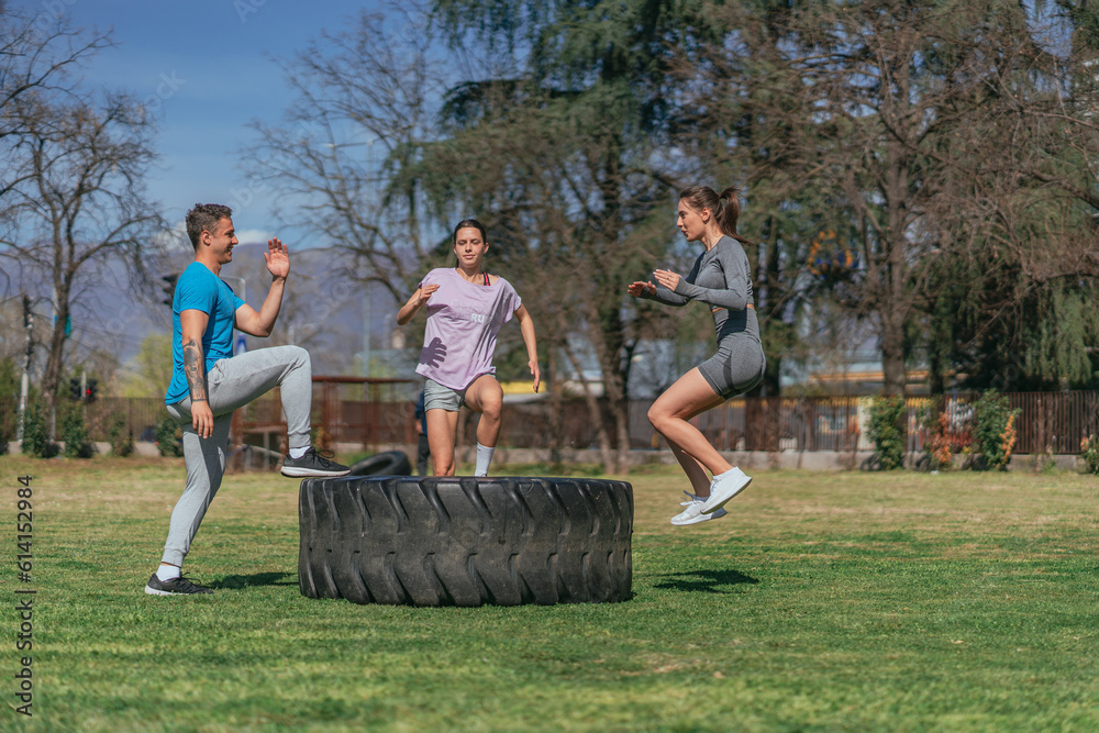 Obraz premium Fit sports friends exercising in the park, on a sunny day. They are doing jumps on a big truck tire