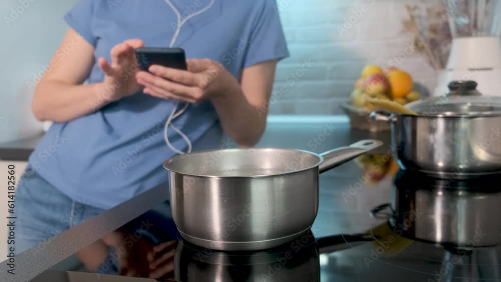 Selective focus on boiling water in saucepan on black induction cooking. Young caucasian woman watching in mobile phone, social media or stories. Modern kitchen with household appliances, cookware. Selective focus on boiling water in saucepan on black induction cooking. Young caucasian woman watching in mobile phone, social media or stories. Modern kitchen with household appliances, cookware.