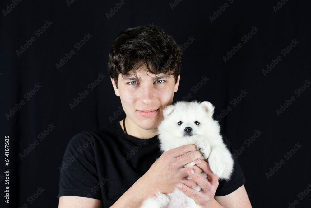 A teenage boy gently holds his little white puppy dog and smiles on dark background, he has long dreamed of such a friend with whom you can walk and talk A teenage boy gently holds his little white puppy dog and smiles on dark background, he has long dreamed of such a friend with whom you can walk and talk