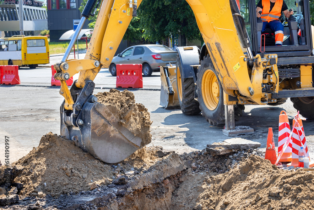 The bucket of a large construction excavator digs a trench on the road against the backdrop of a city street on a sunny summer day.