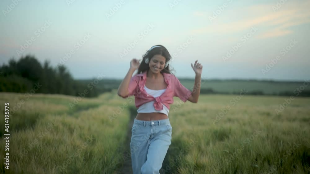 Young beautiful woman in jeans and checkered shirt listens to music and dances while walking in the field during sunset Young beautiful woman in jeans and checkered shirt listens to music and dances while walking in the field during sunset