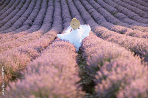 Happy woman in a white dress and straw hat strolling through a lavender field at sunrise, taking in the tranquil atmosphere.
