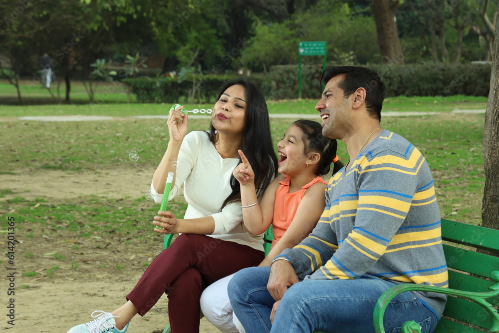 Parents enjoying with daughter in park surrounded with greenery and serenity. They are having joyful and cheerful time together on a bench in lush green environment. Loads of smile and happy moments.