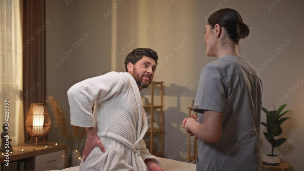 Handsome Caucasian male patient having back issues while sitting on bed in wellness center. Young female massagist consulting or recommending exercises which renovate spinal muscles. Handsome Caucasian male patient having back issues while sitting on bed in wellness center. Young female massagist consulting or recommending exercises which renovate spinal muscles.