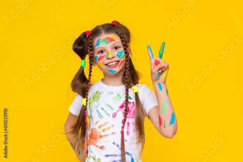 A young girl stained in multicolored paint shows two fingers and smiles broadly. Children's creativity. Yellow isolated background.