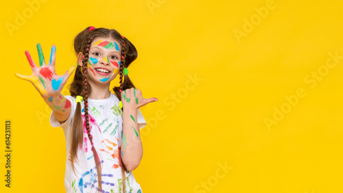 A young girl stained in multicolored paint shows five fingers and smiles broadly pointing at your advertisement. Children's creativity. Copy space. Yellow isolated background.