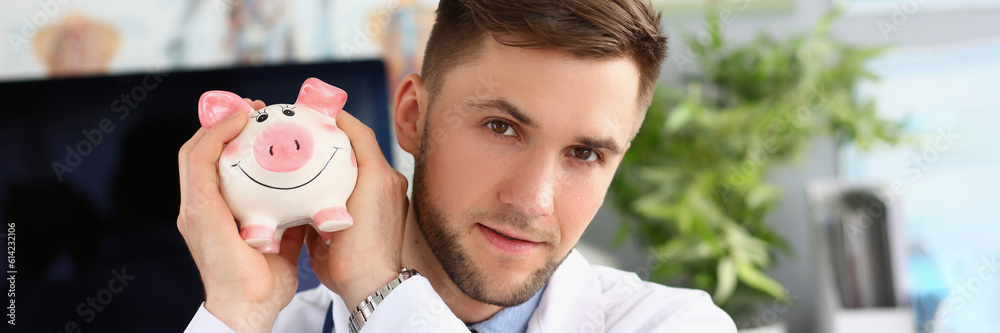 Fototapeta premium Portrait of male doctor holding a piggy bank on voluntary donations of money for a hospital or clinic. General practitioner collects charitable donation for patient care healthcare concept
