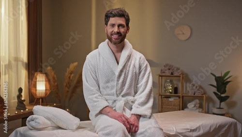 Positive Caucasian man having resting day on his weekend in beauty salon. Bearded male with tore sitting on message bed and cherfully smiling on camera. Wellness and relaxation services. photo