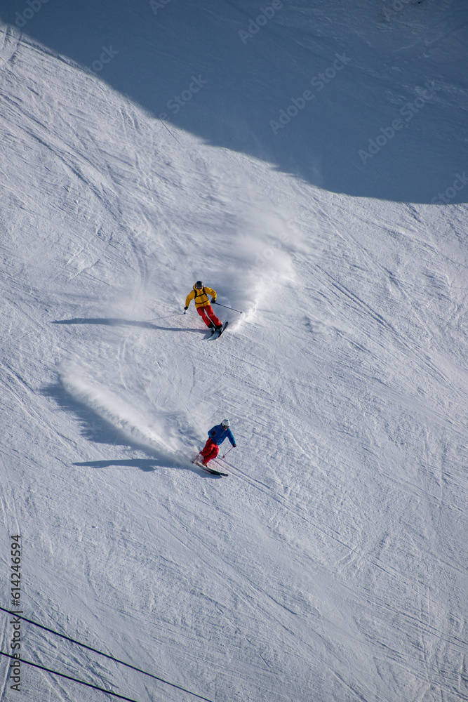 skiing down mountain slope with white snow powder projecting from behind, skiers wearing black and blue  and yellow and orange outfit, Les Deux Alpes, Isère, Auvergne-Rhône-Alpes, France, 2/2
