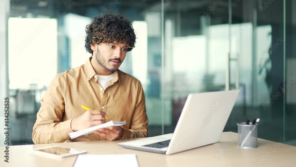 A young employee watching video call training notes in a notebook looking at the laptop screen sitting at workplace in office. Man is studying online, listening remotely to a course, webinar, seminar A young employee watching video call training notes in a notebook looking at the laptop screen sitting at workplace in office. Man is studying online, listening remotely to a course, webinar, seminar