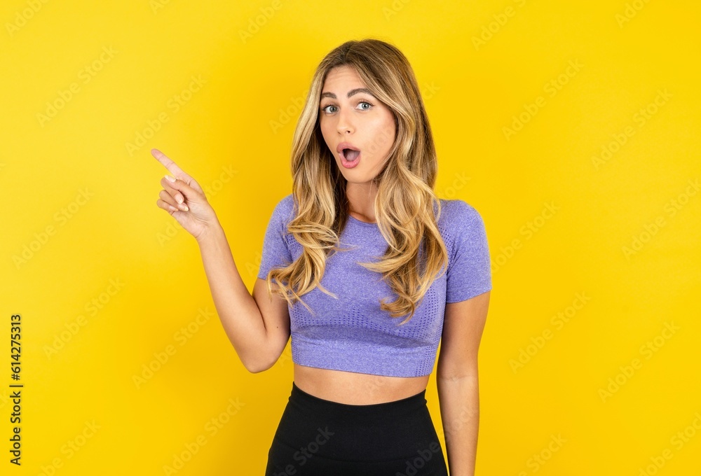 Emotive young beautiful blonde woman wearing sportswear over yellow stukeeps jaw dropped from shock demonstrates amazing promo points right on blank space demonstrates big shopping sale. Advertisement Emotive young beautiful blonde woman wearing sportswear over yellow stukeeps jaw dropped from shock demonstrates amazing promo points right on blank space demonstrates big shopping sale. Advertisement
