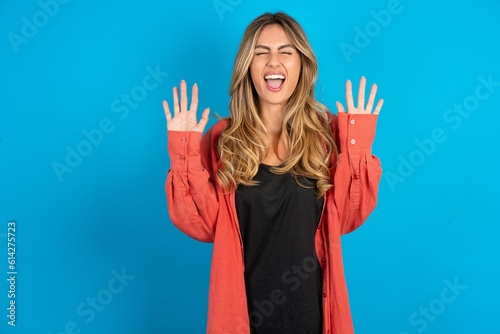 Emotive young beautiful blonde woman standing over blue studio background  laughs loudly  hears funny joke or story  raises palms with satisfaction 