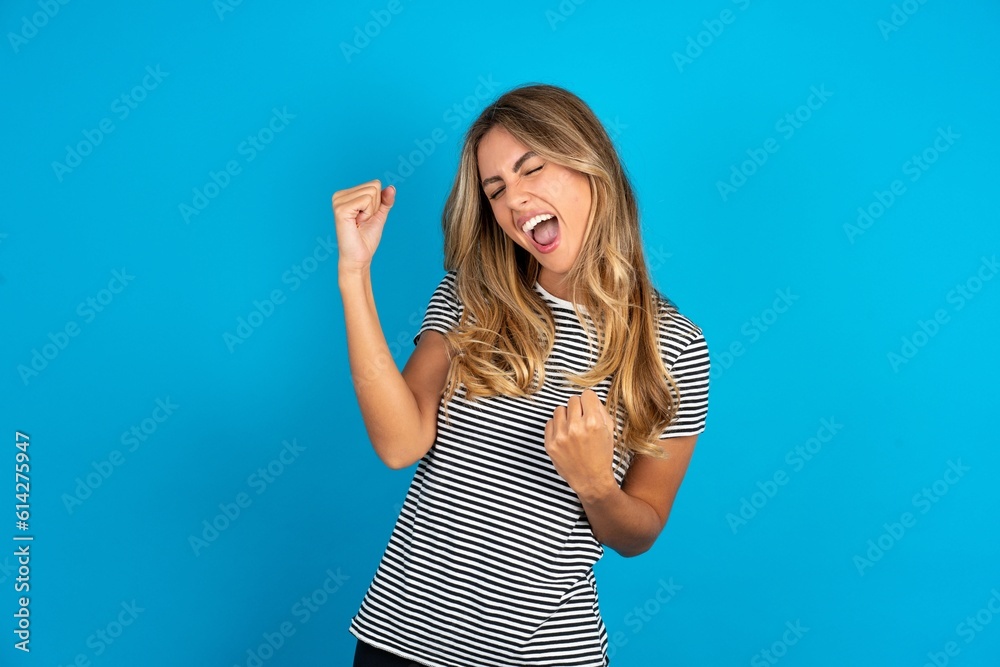 Young beautiful blonde woman wearing striped t-shirt over blue studio background very happy and excited doing winner gesture with arms raised, smiling and screaming for success. Celebration concept. Young beautiful blonde woman wearing striped t-shirt over blue studio background very happy and excited doing winner gesture with arms raised, smiling and screaming for success. Celebration concept.