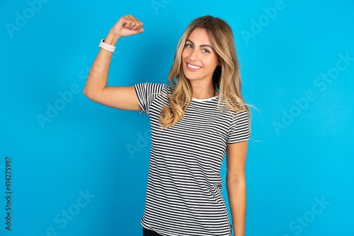 Portrait of powerful cheerful Young beautiful blonde woman wearing striped t-shirt over blue studio background showing muscles.