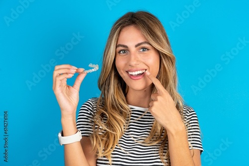 Young beautiful blonde woman wearing striped t-shirt over blue studio background holding an invisible aligner and pointing to her perfect straight teeth. Dental healthcare and confidence concept.